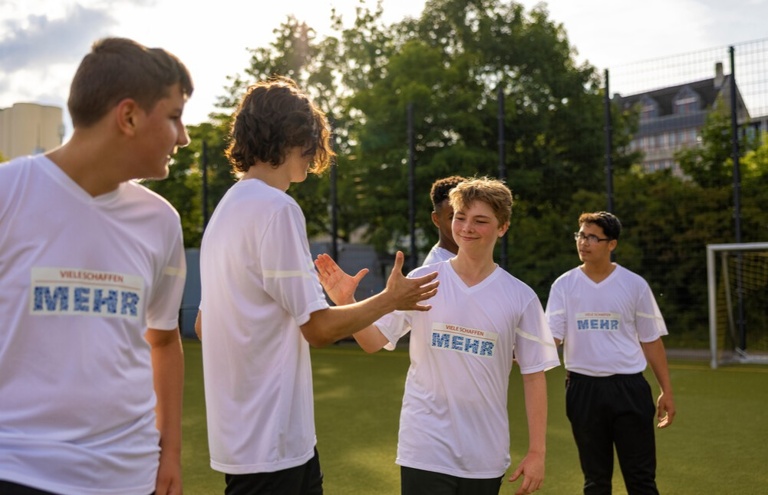 Mehrere Jugendliche in "Viele schaffe mehr"-Sporttrikots stehen auf einem Fußballplatz. Zwei geben sich ein High-Five, andere schauen lächelnd zu. Im Hintergrund sind Tore, Bäume und Gebäude zu sehen